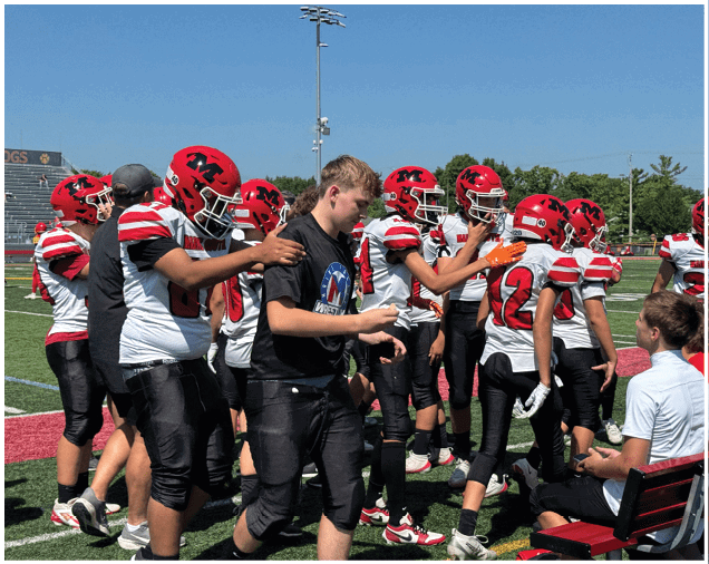 A group of people wearing red helmets and white and red striped football uniforms stand on a field while one person in black sportswear interacts with them near a bench where two people are seated. Description generated by AI
