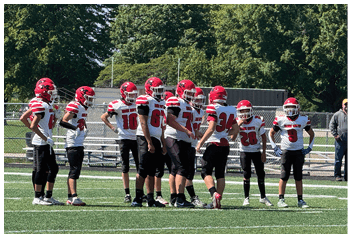 A group of people wearing red helmets and white sports uniforms with red stripes are standing outdoors near a fence with trees in the background. Description generated by AI