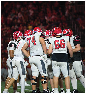 A group of football players in white and black uniforms with red helmets are huddled together on a field during a game. Description generated by AI