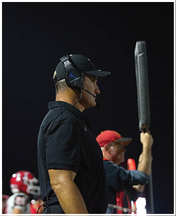 A person wearing a black polo shirt and a headset stands on the right side of the image with a dark background and blurred red helmets visible in the lower left corner. Description generated by AI