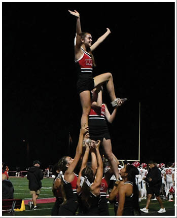 A person wearing a red and black cheerleading uniform with arms raised and smiling against a dark background. Description generated by AI