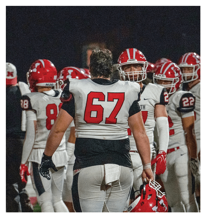 A group of people wearing football uniforms and helmets are gathered closely together on a sports field, appearing to be in a team huddle or discussion. Description generated by AI