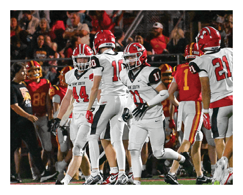 A person wearing a red helmet and black football uniform with the number 14 stands on a football field while other players and a referee are visible in the background. Description generated by AI
