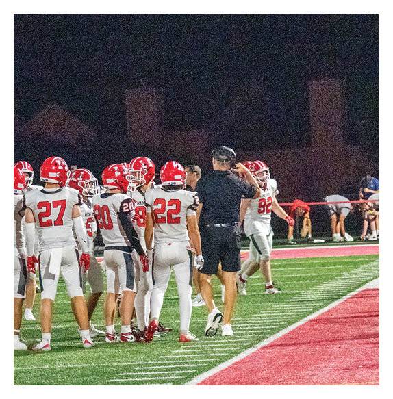 A group of people wearing red helmets and black or white sports uniforms are engaged in a football play on a green field with trees and a fence in the background. Description generated by AI