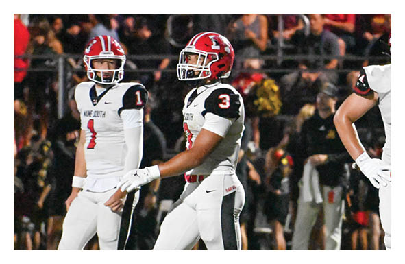 Two people wearing black sports uniforms and red helmets are positioned on a football field with trees and a fence in the background. Description generated by AI