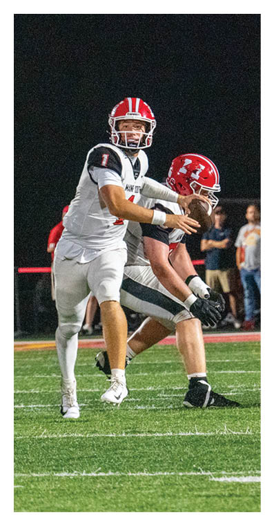 Two people wearing red helmets and black football uniforms are on a football field, with one person preparing to throw a football and the other in a blocking stance. Description generated by AI