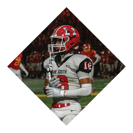 Two people wearing red helmets and sports uniforms are on a football field with one person holding the ball on the ground while the other is kicking it. Description generated by AI