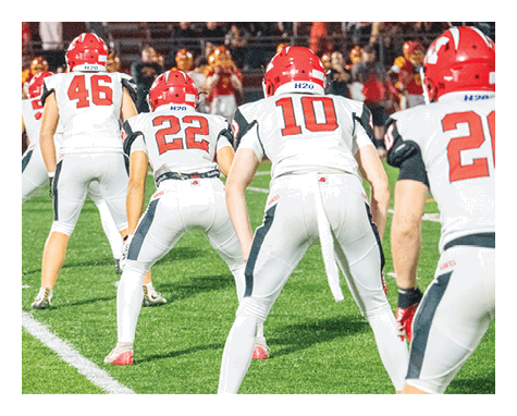 People wearing helmets and sports uniforms are playing football on a field with bleachers in the background. Description generated by AI