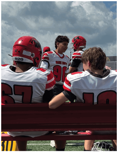 People in football uniforms are sitting and standing on the sidelines of a football field under a partly cloudy sky. Description generated by AI
