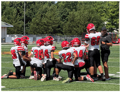A group of people wearing red helmets and white sports jerseys are kneeling on a football field while one person in black sportswear stands and talks to them. Description generated by AI