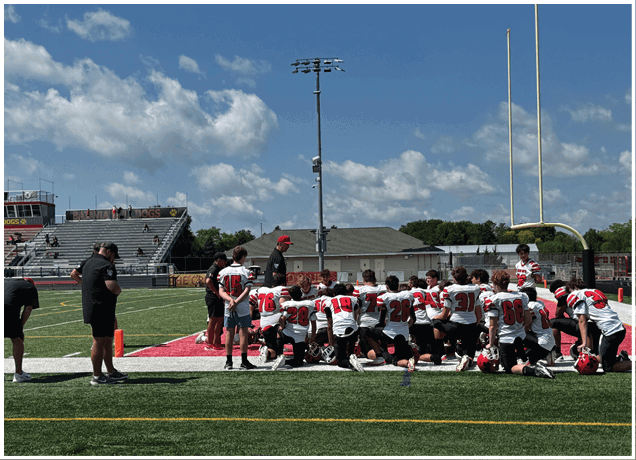 A group of people wearing football uniforms and helmets are gathered on a field near a goalpost while a few others stand nearby on a sunny day. Description generated by AI