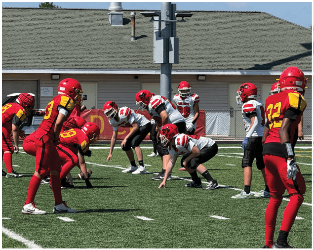 People in red and white football uniforms are lined up on a green field preparing for a play with a building and blue sky in the background. Description generated by AI