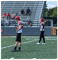 Two people in football uniforms with red helmets stand on a green field while several people sit in the bleachers and on benches in the background. Description generated by AI