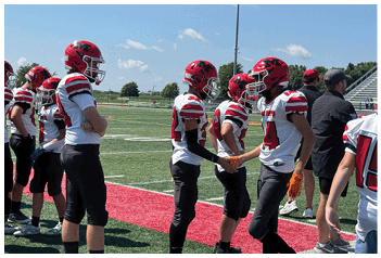 Several people wearing red helmets and white football jerseys are standing on a sports field, with two of them shaking hands. Description generated by AI
