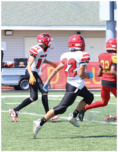 Four people in football uniforms are actively playing on a field with one person running, two standing, and one person crouched down near another person on the ground. Description generated by AI