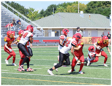 Adults in football uniforms are playing a game on a green field with bleachers and a building in the background. Description generated by AI