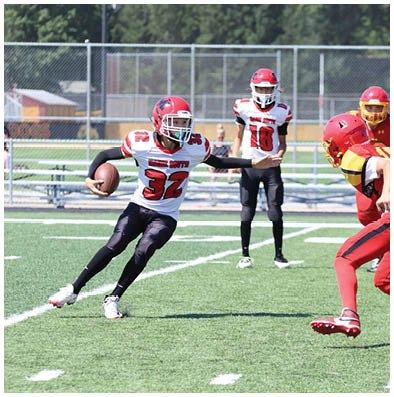 People in football uniforms are actively playing on a green field with one person running with the ball while others are preparing to block or tackle. Description generated by AI