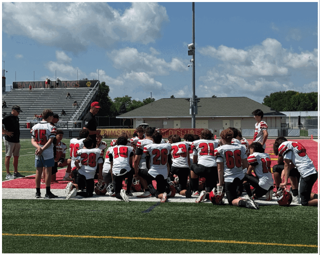 A group of people wearing white and red football uniforms are gathered on a green field with some standing and others kneeling while two adults in casual clothing stand nearby. Description generated by AI