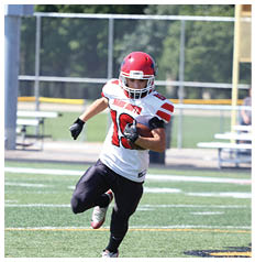 An adult wearing a red helmet and white football jersey is running on a grassy field while holding a football. Description generated by AI