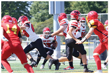 Players in red and white football uniforms are actively engaged in a play on a grassy field with trees and bleachers in the background. Description generated by AI