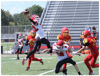 Players in red and white football uniforms are actively engaged in a play on a green field with empty bleachers in the background. Description generated by AI