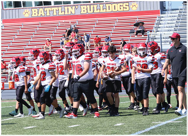 A group of people wearing football uniforms and helmets are walking on a football field near bleachers with a sign that reads “BATTLIN' BULLDOGS." Description generated by AI