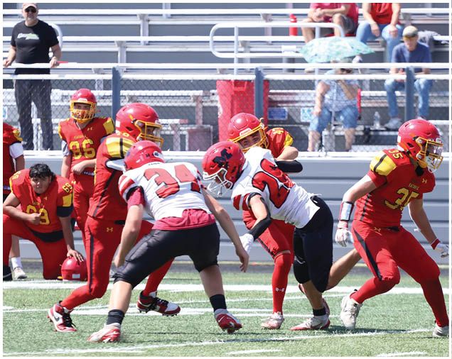 Players in red and white football uniforms are actively engaged in a play on a green field with spectators seated in bleachers behind them. Description generated by AI
