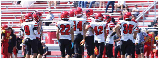 A group of football players in red helmets and white jerseys with black pants are gathered on a field with empty bleachers and scattered spectators in the background. Description generated by AI