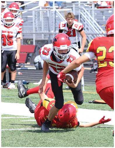 People in football uniforms are actively playing on a field with one person running with the ball while another is on the ground attempting a tackle. Description generated by AI