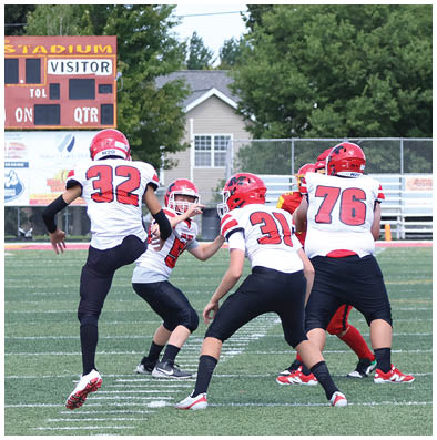 Adults in football uniforms are engaged in a game on a green field with a scoreboard and bleachers in the background. Description generated by AI