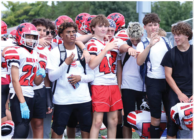 A group of young people wearing football uniforms and helmets stand together outdoors, some holding helmets and others with backpacks. Description generated by AI