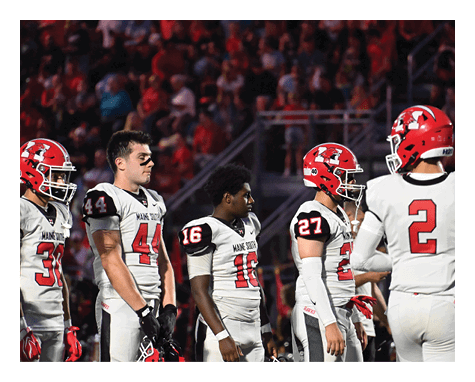 People wearing white and red football uniforms stand together on a field, some holding helmets. Description generated by AI