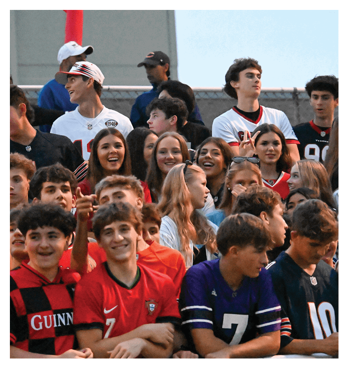 A group of people wearing various sports jerseys are gathered closely together, smiling and looking in different directions, with a chain-link fence in the background. Description generated by AI