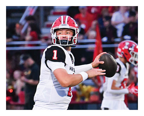 A person wearing a white football uniform with a red helmet is preparing to throw a football on a field with other players and spectators in the background. Description generated by AI