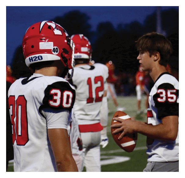 Three people in football uniforms stand on a field at dusk, with one holding a football and the others facing away. Description generated by AI