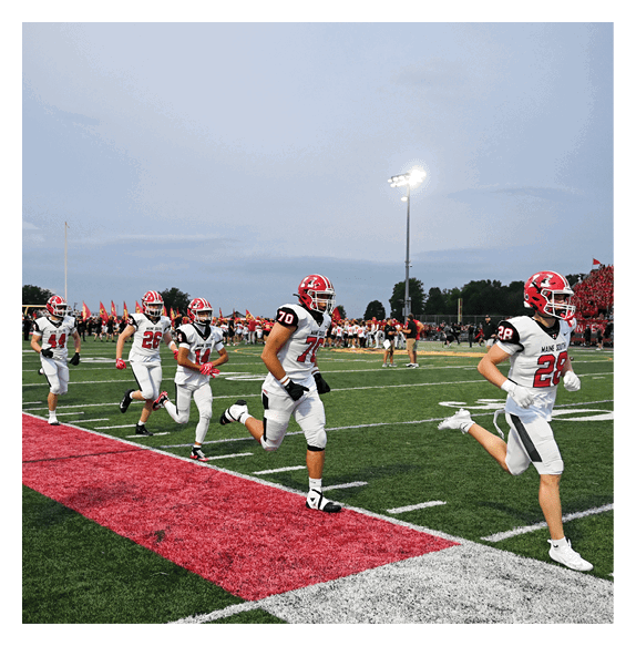 A group of people wearing white football uniforms with red helmets are running onto a green football field with a red and white sideline, while a crowd watches from the stands in the background. Description generated by AI
