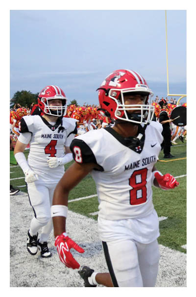 Two people in football uniforms are running on a field with a marching band playing instruments in the background. Description generated by AI