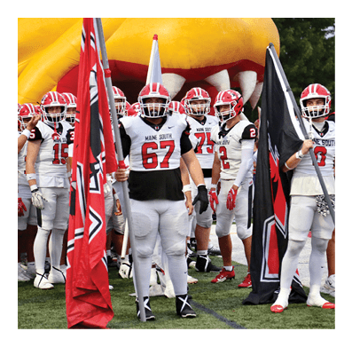 Several people in football uniforms holding flags stand on a field with a large inflatable mascot in the background. Description generated by AI