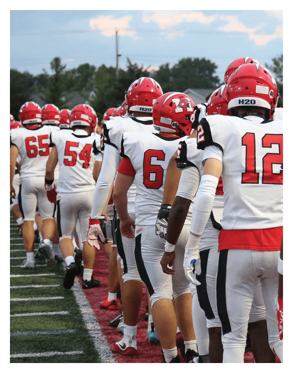 A line of people wearing red helmets and white sports uniforms stand on a football field, shaking hands with others. Description generated by AI