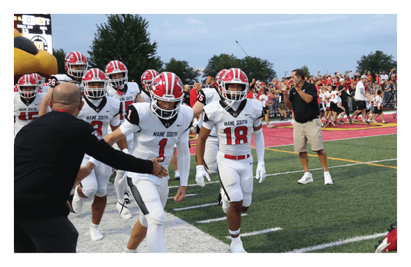 A crowd of people is gathered outdoors near a football field with some wearing sports jerseys and a person in a red football helmet visible on the right side. Description generated by AI