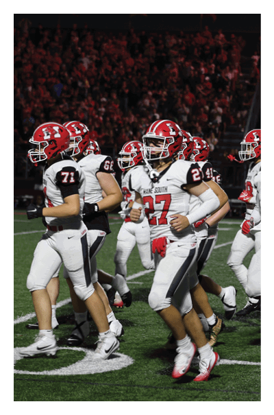 A group of people wearing white football uniforms with red helmets are running on a green field during a nighttime game. Description generated by AI
