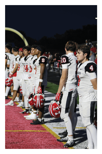 A line of people in white and black football uniforms stand side by side on a red and white field under stadium lights at night. Description generated by AI