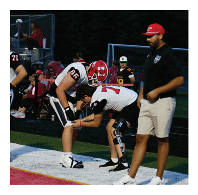 Three people in football uniforms and one person in casual sportswear stand on the sideline of a football field, with two players interacting closely and a coach observing nearby. Description generated by AI