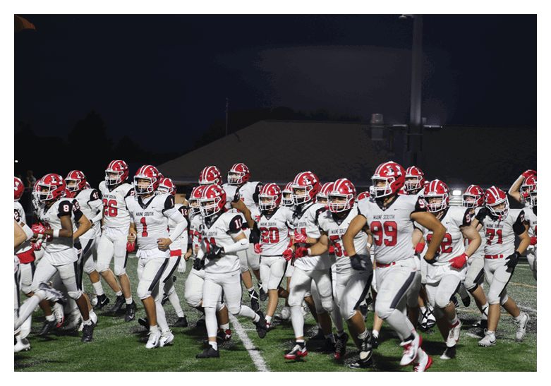 A group of people wearing white sports uniforms with red and black accents are running together on a green field marked with white lines. Description generated by AI