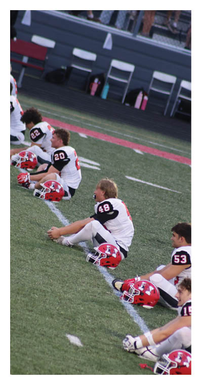 Several people in football uniforms are sitting on the grass field stretching with their helmets placed in front of them. Description generated by AI