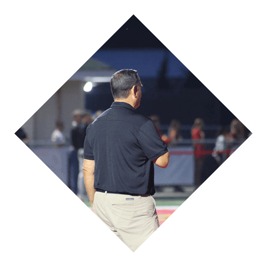 A person wearing a black shirt and beige pants stands on a sports field with a red and yellow surface, facing away from the camera. Description generated by AI