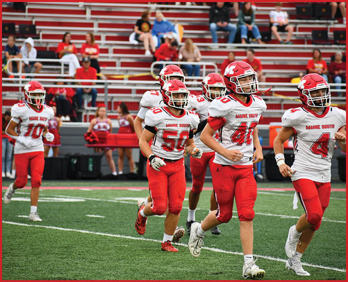 Several people wearing red and white football uniforms are walking on a green field with bleachers and spectators in the background. Description generated by AI