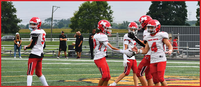 Several people wearing red and white football uniforms are standing and interacting on a green outdoor sports field with trees and spectators in the background. Description generated by AI