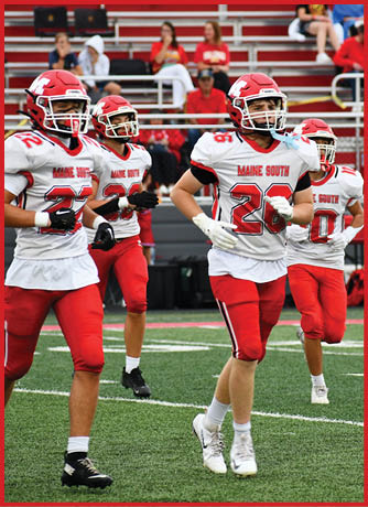 Four people in red and white football uniforms are jogging on a green field with a red and white stadium background. Description generated by AI