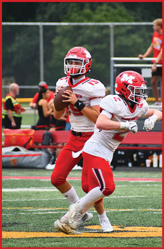 Two football players in red and white uniforms are on a field with one holding a football ready to throw and the other running ahead. Description generated by AI
